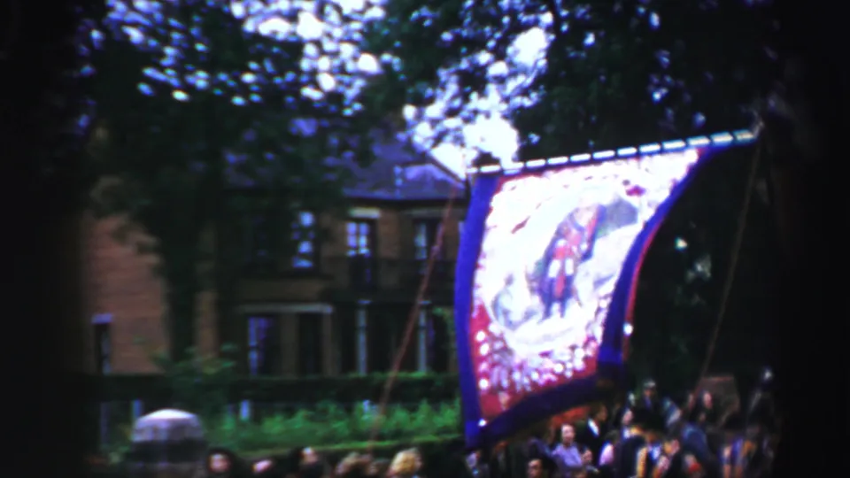 A decorated banner passes through a tree-lined United Kingdom parade route, with a building partly visible behind it.
