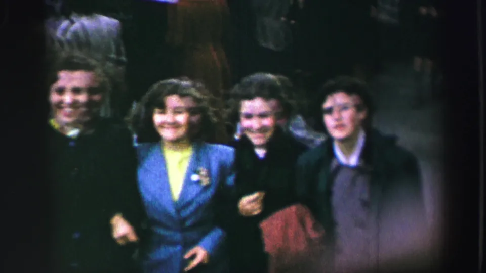 Four women in 1950s-style coats and dresses walk arm in arm toward the camera during a United Kingdom parade scene.