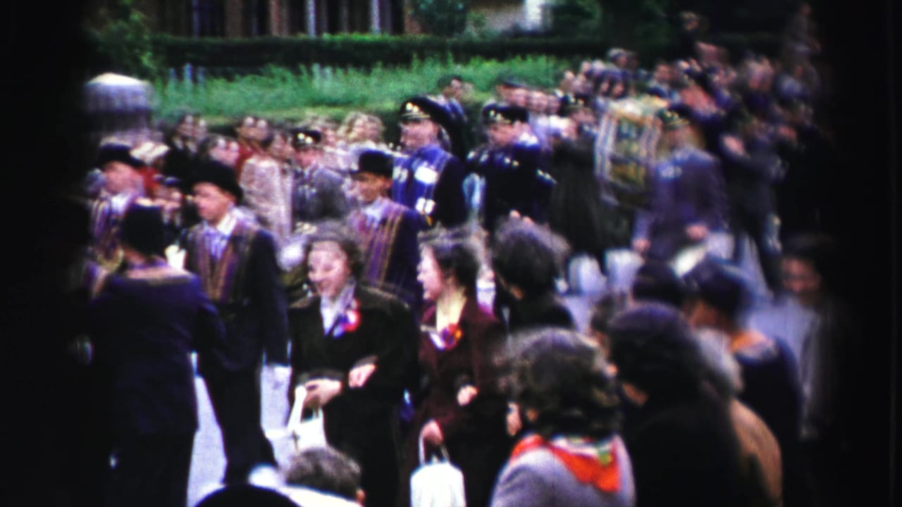 Marchers in dark formal clothing and sashes pass spectators on a street.