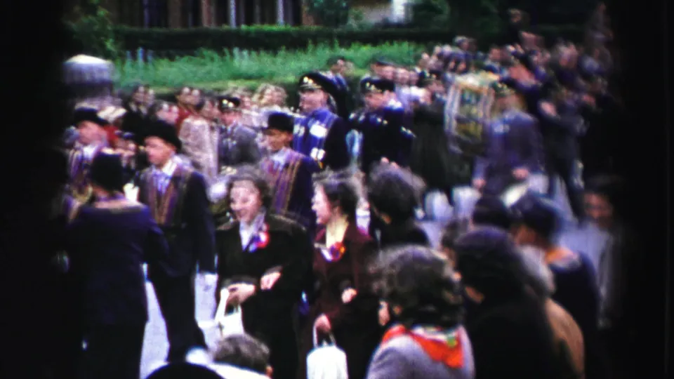 1952 United Kingdom home-movie frame of formally dressed marchers with bowler hats and sashes moving past a sidewalk crowd.