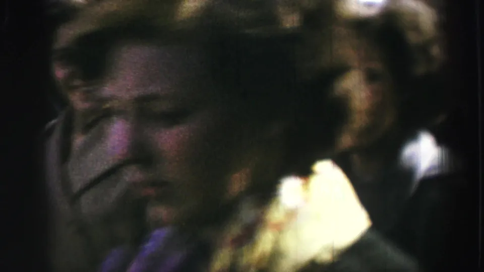 Soft 8mm frame from a United Kingdom public event showing adults close to the camera, with faces partly visible in the crowd.
