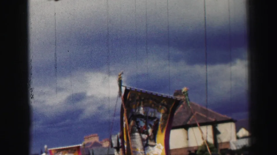 The 8mm frame shows the parade banner passing near rooftops, with a large cloudy sky and visible edge shadows from the film scan.