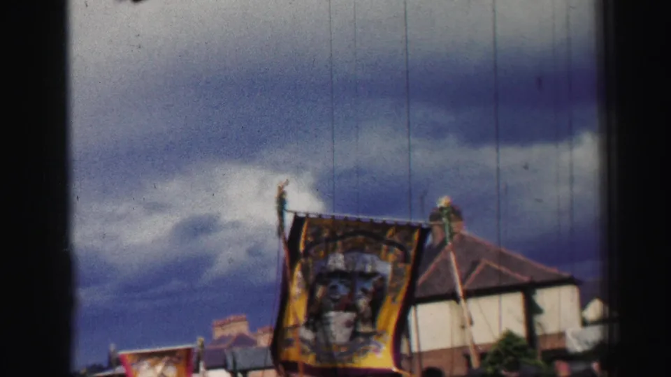 A colorful religious banner is carried through the United Kingdom street scene, with buildings, poles, and heavy clouds visible in the frame.