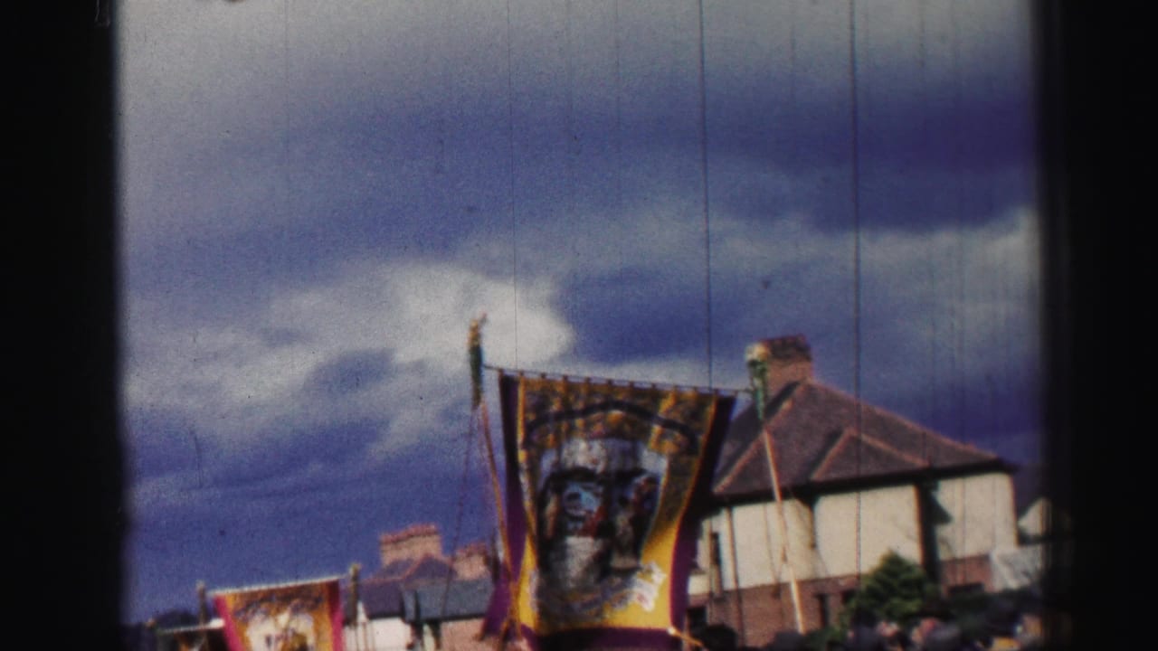 1952 United Kingdom home-movie frame showing an ornate parade banner, residential houses, and spectators under a dramatic sky.