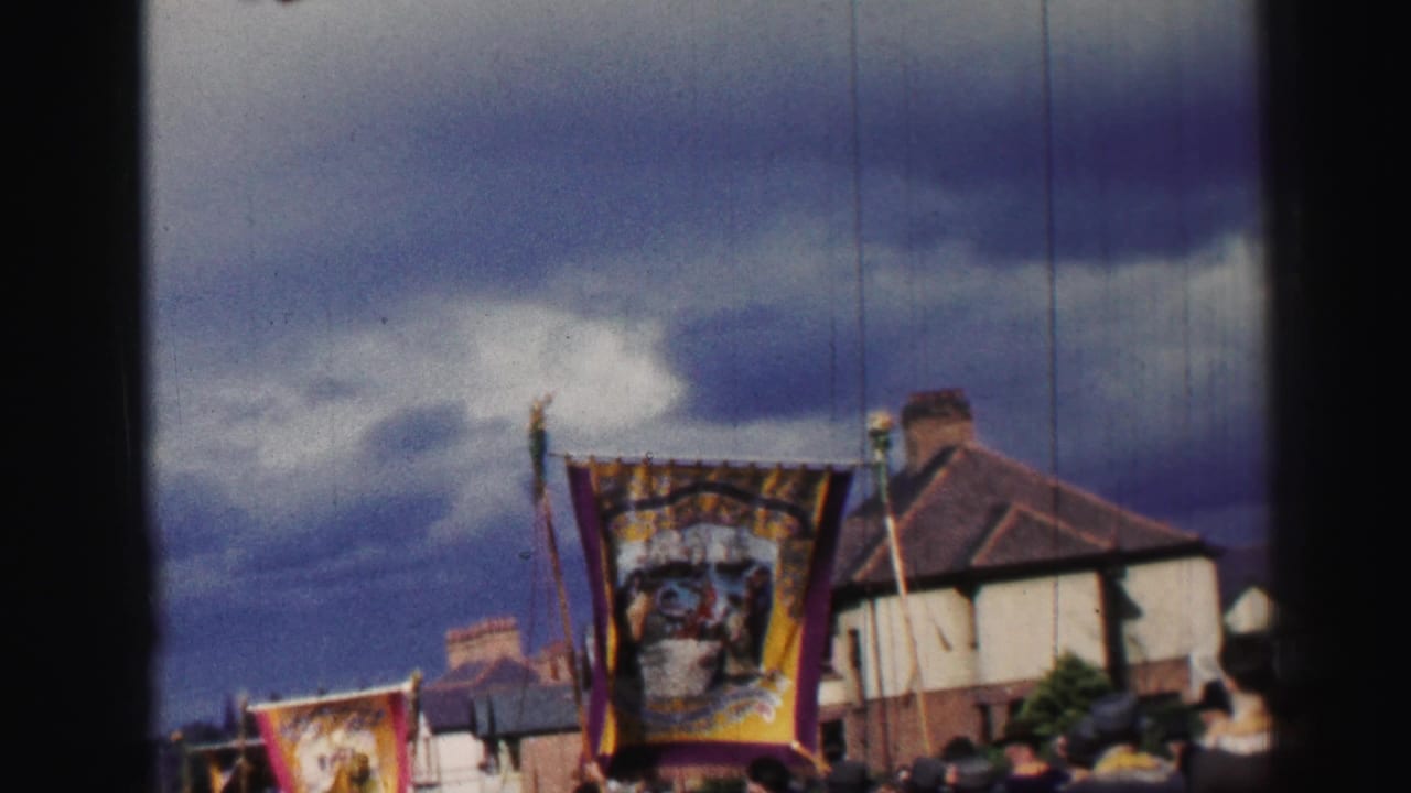 A large ornate banner is carried through a British residential street as spectators watch from below the frame.