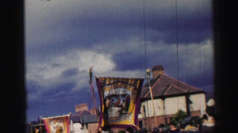 A large ornate banner is carried through a British residential street as spectators watch from below the frame.