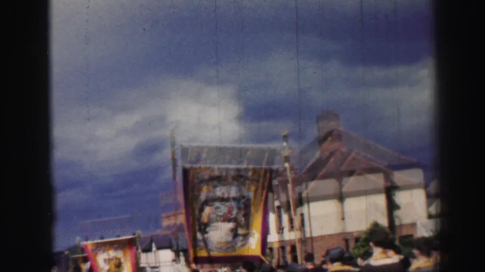 Soft 8mm frame of the procession with a carried banner, house fronts, and onlookers along the street.