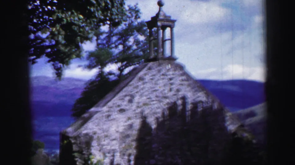 A 1952 United Kingdom 8mm view shows a stone church roofline with a small bell tower, trees, cloudy sky, and distant hills.