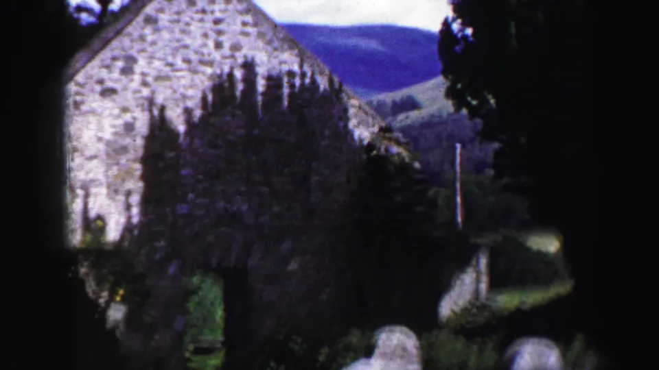 1952 United Kingdom 8mm frame showing a weathered stone building seen through trees, with hillside visible beyond.