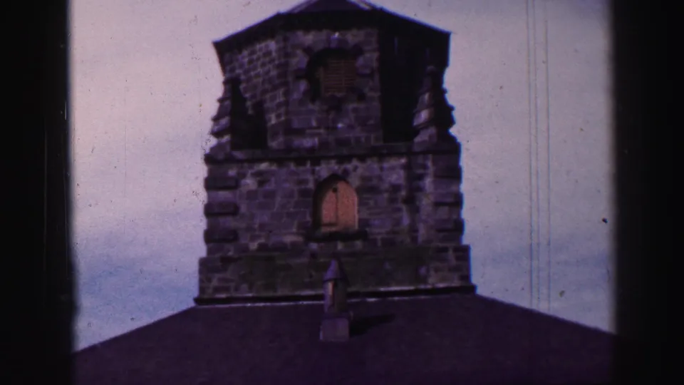 1952 United Kingdom home-movie frame showing the upper part of a weathered stone tower above a dark roofline.