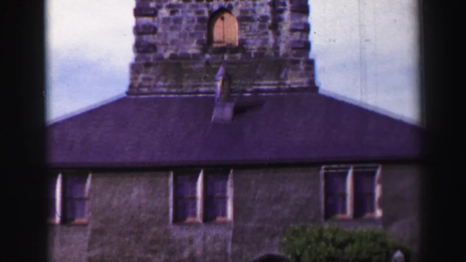 Mid-tilt frame showing the historic building facade with windows, shrubs, dark roof, and tower above.
