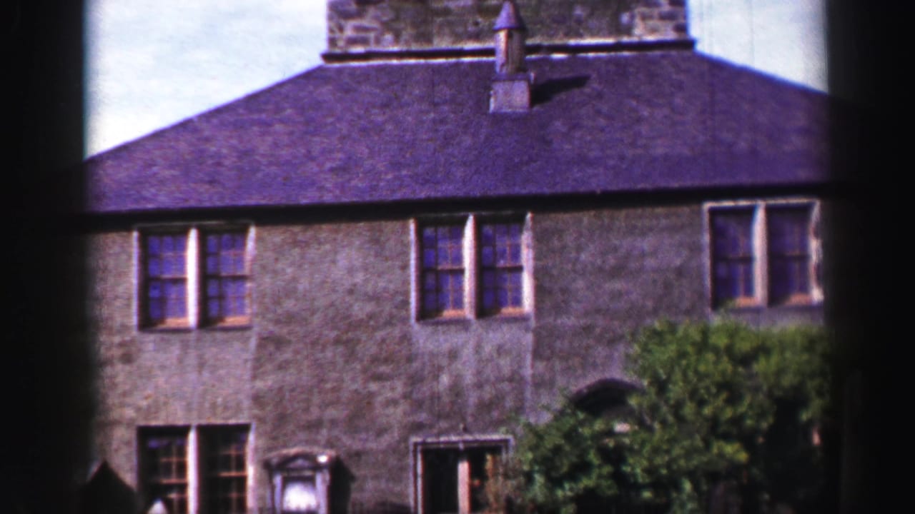 1952 United Kingdom 8mm frame showing an old stone building facade with shuttered windows, a dark roof, chimney and leafy growth.