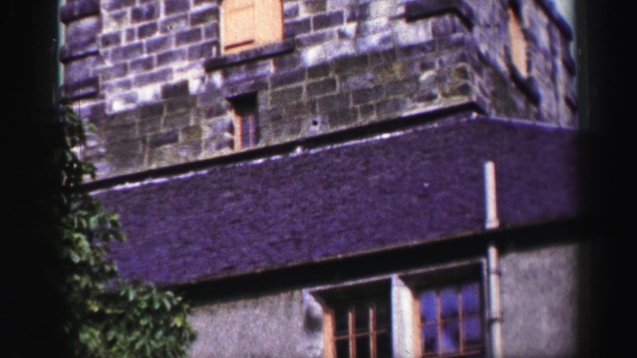 1952 United Kingdom 8mm frame of a weathered stone corner, dark purple-toned roof edge, and tree foliage beside the facade.