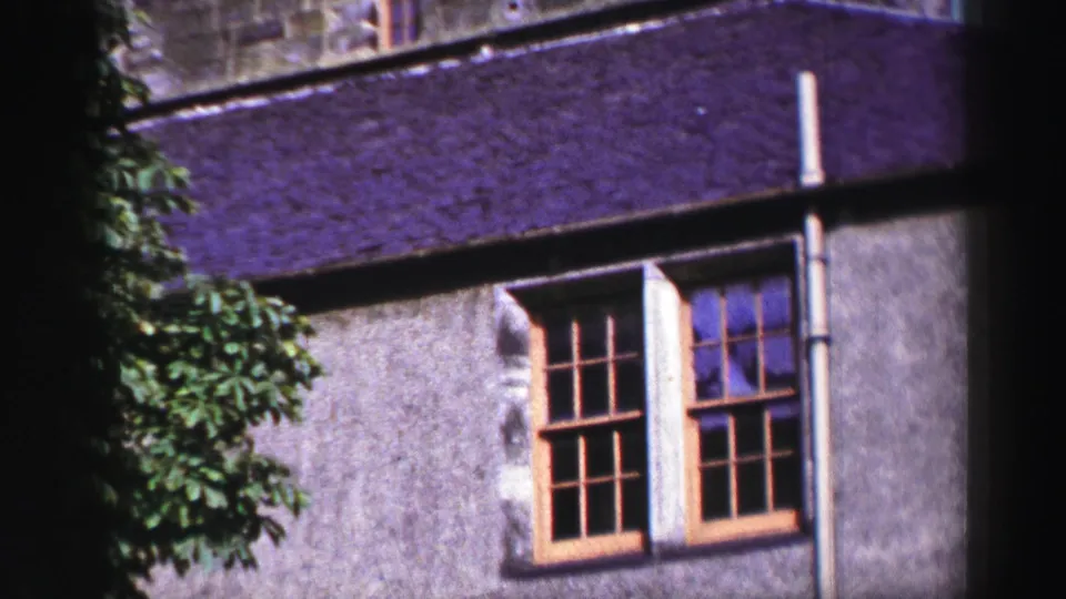 1952 United Kingdom 8mm frame showing a lower exterior wall with multi-pane wooden windows, purple-toned roof surface, and tree leaves.