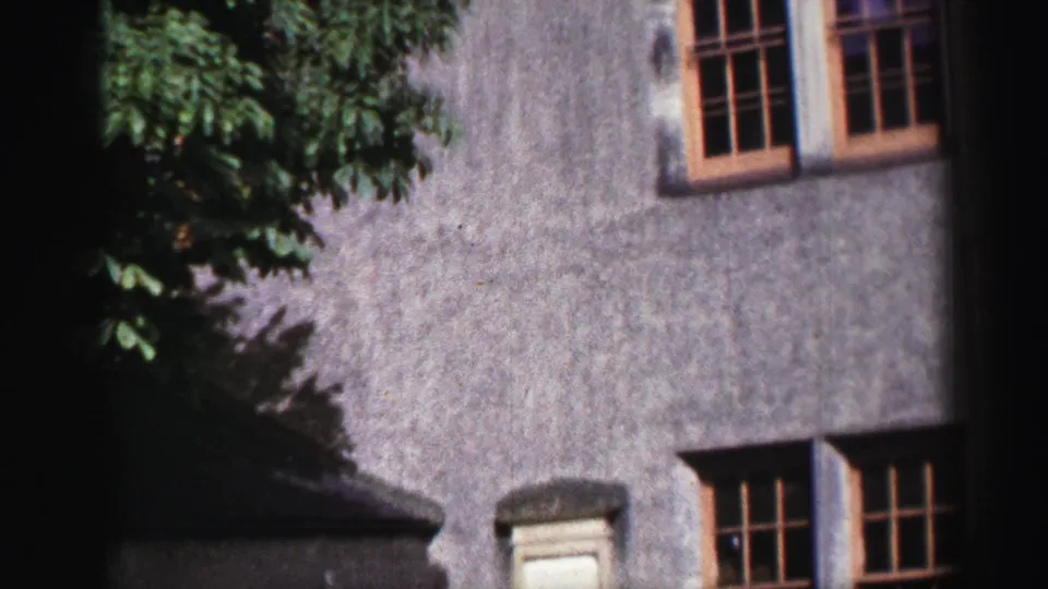 1952 United Kingdom 8mm frame of the building's gray lower wall, wooden multi-pane windows, roof shadow, and upper stonework.