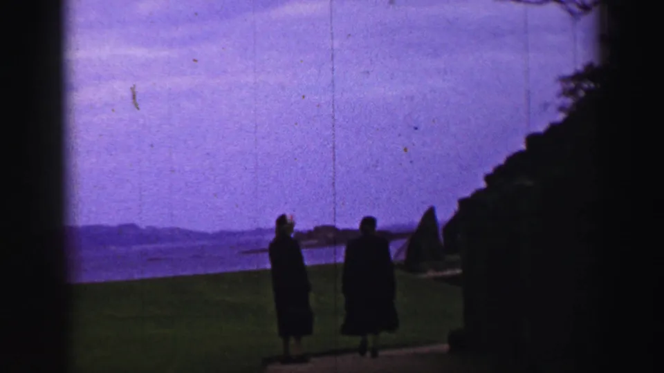 Two dark-clad visitors are seen on the grass near Inchcolm Abbey with the water and distant shore in the background.
