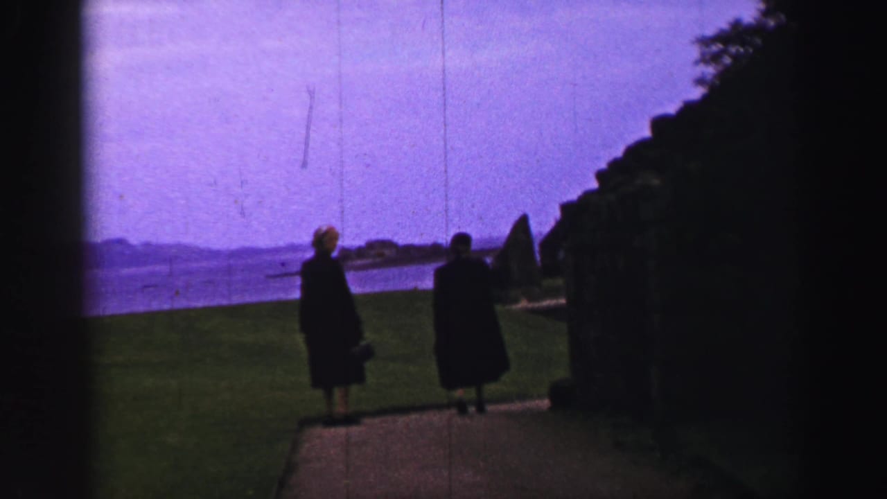 Two women in 1952 United Kingdom home-movie footage stand near a coastal path, with bay water and a dark hedge beside them.