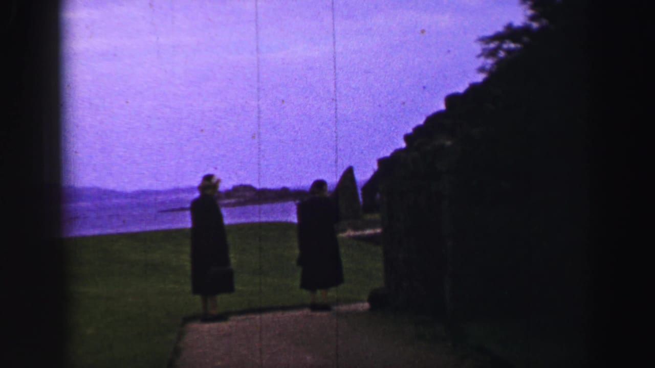 Two women in dark coats stand near a hedge on a United Kingdom coastal path, looking across the bay in soft 8mm color.