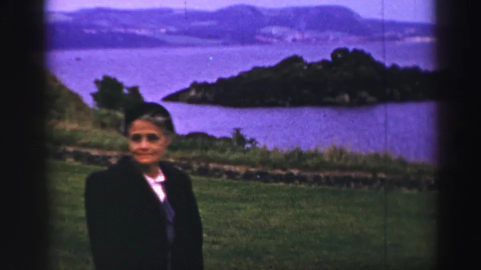 A woman in formal 1950s-style clothing faces the camera in a United Kingdom coastal scene with a bay, island, and hills behind her.