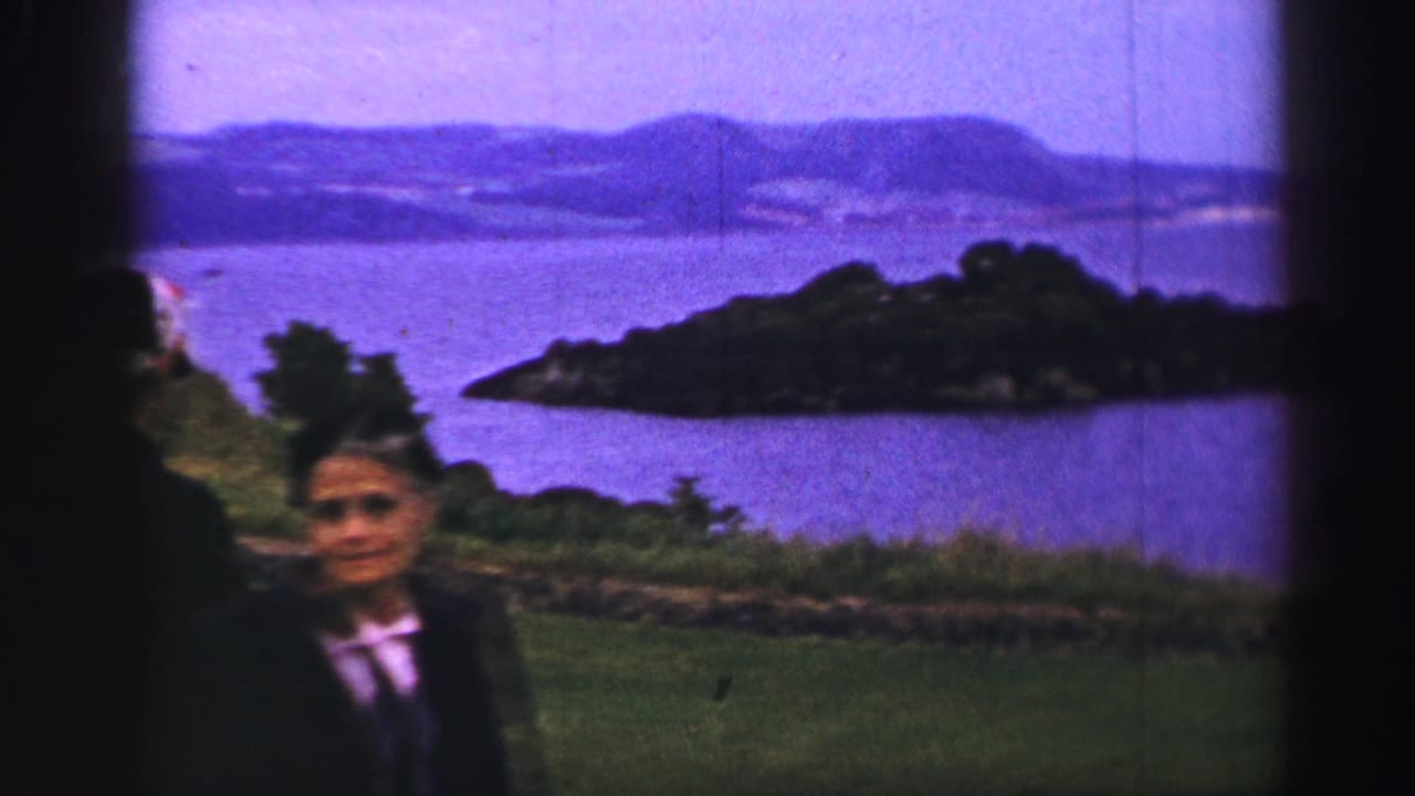 1952 United Kingdom home-movie frame showing a woman in dark clothing on a grassy overlook above water and a tree-covered island.