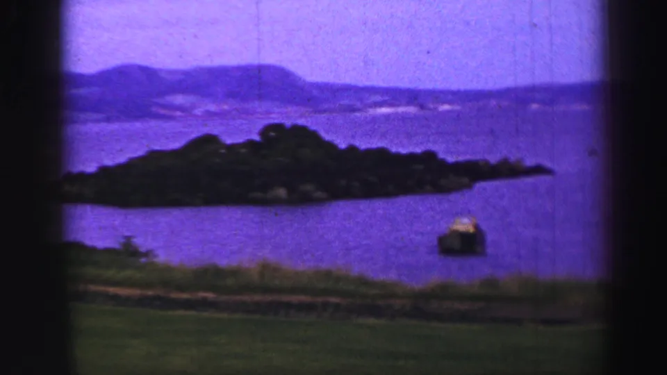1952 United Kingdom 8mm frame centered on a small wooded island across water, with distant rolling hills and shoreline grass.