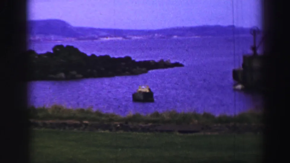 1952 United Kingdom home-movie frame showing a small boat on water, a wooded island, distant hills, grass, and part of a dock structure.