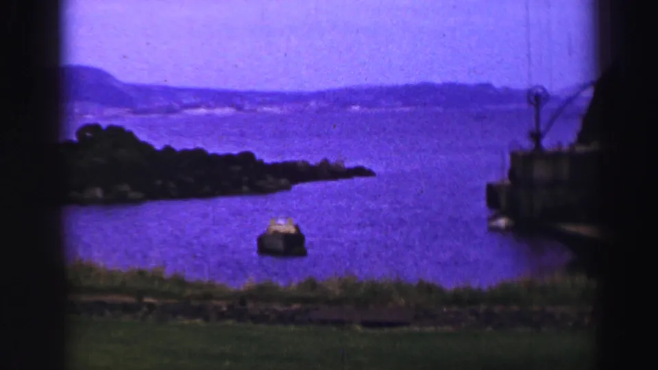 1952 United Kingdom 8mm frame with a small boat on blue-purple water, a wooded island to the left, and a dock structure at right.