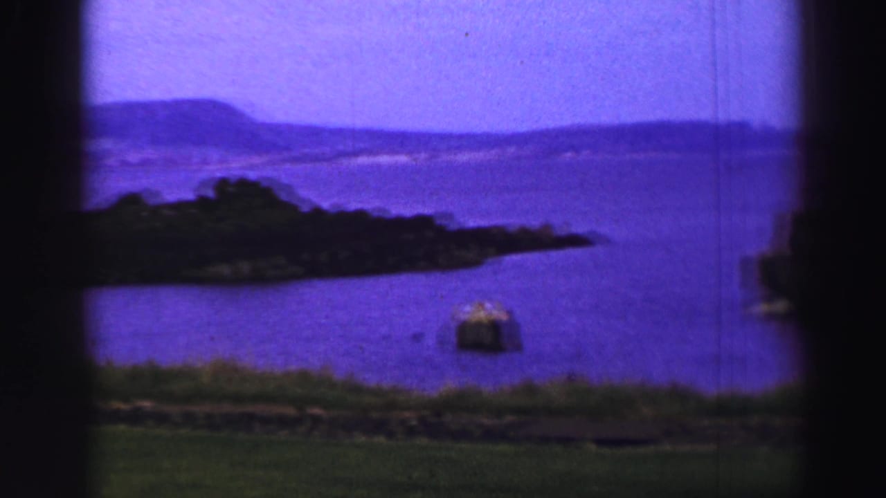 1952 United Kingdom 8mm frame showing purple-tinted coastal water, a small island, grassy foreground, and distant hills.