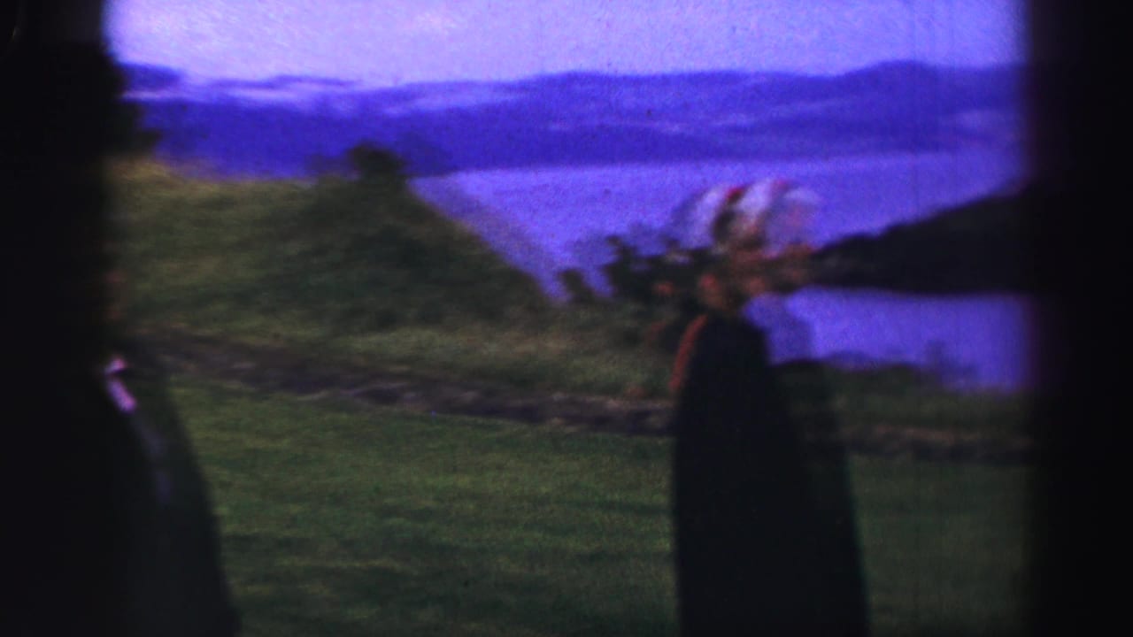 Person in dark clothing standing on grass and looking toward water and distant hills.