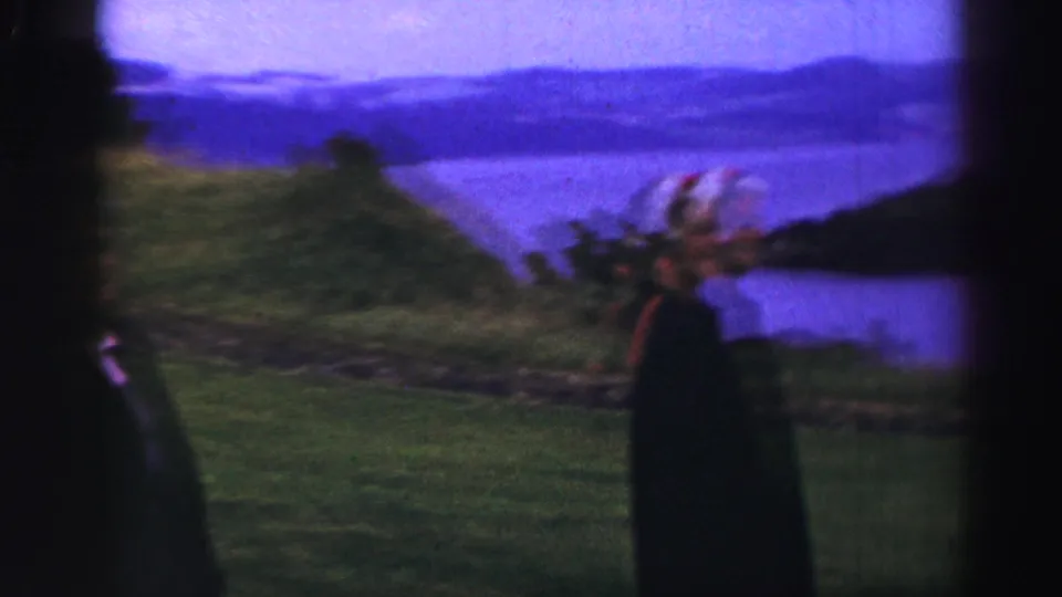 A visitor in dark period clothing stands on a grassy rise overlooking water and hills in 1952 United Kingdom home-movie footage.