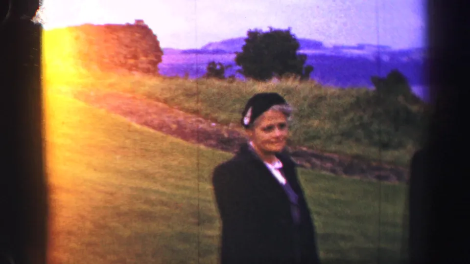 A woman in a dark coat and headscarf stands near castle grounds with coastal water and hills behind her in 1952 UK footage.