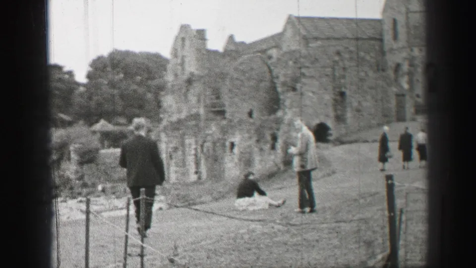 A 1952 United Kingdom tourist scene shows visitors walking along a roped path beside weathered ruin walls.