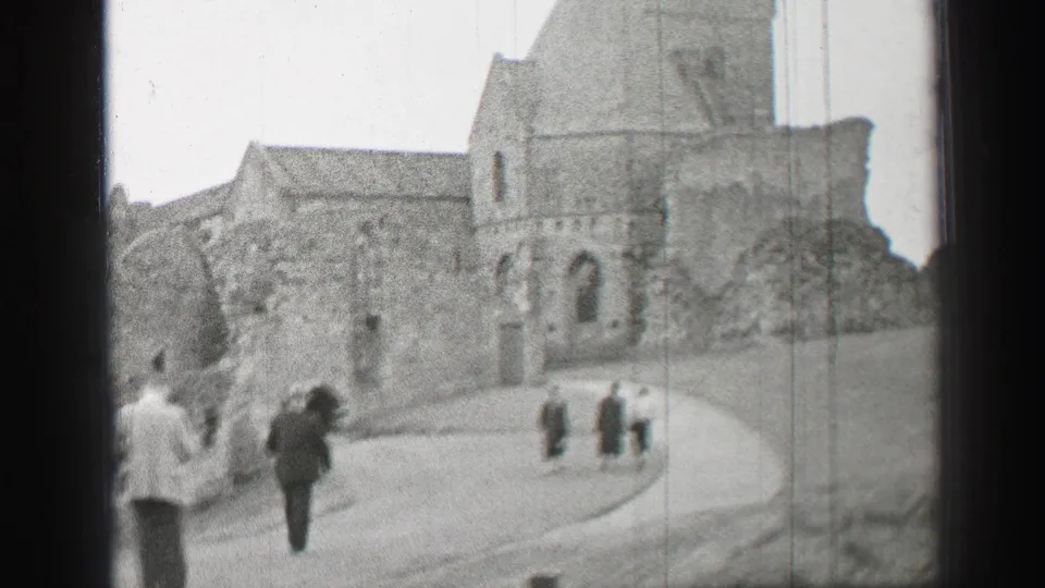 Visitors in formal coats walk uphill toward historic stone church ruins in the United Kingdom, filmed on 8mm in 1952.
