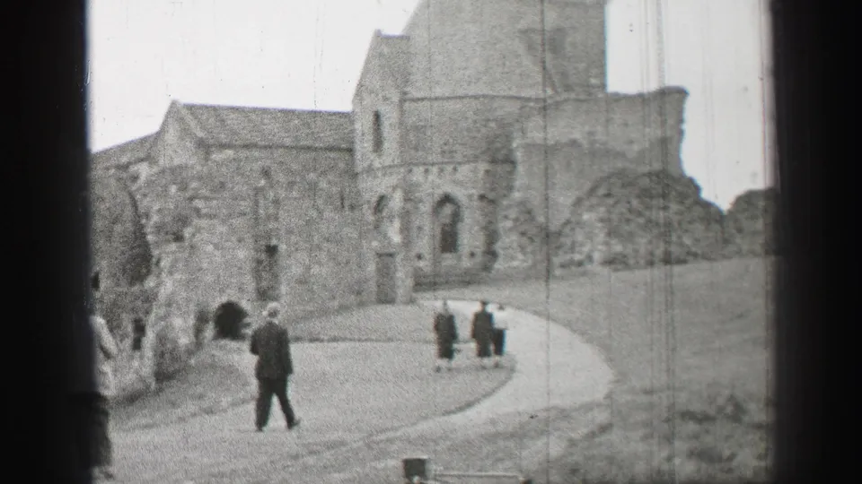 A man in a dark coat crosses in front of stone church ruins while other visitors walk uphill in 1952 home-movie footage.