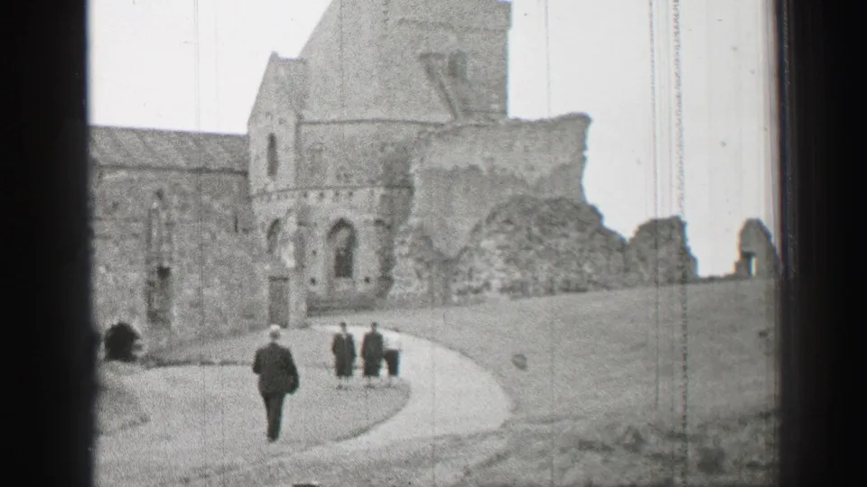 1952 United Kingdom home-movie frame showing formally dressed visitors approaching large stone castle ruins along a curved path.