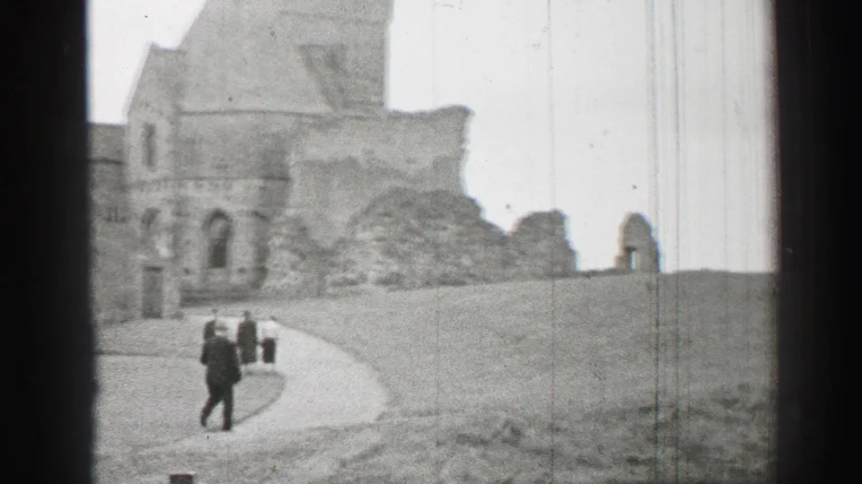 1952 United Kingdom 8mm frame with a formally dressed visitor walking near castle ruins and a curved path.