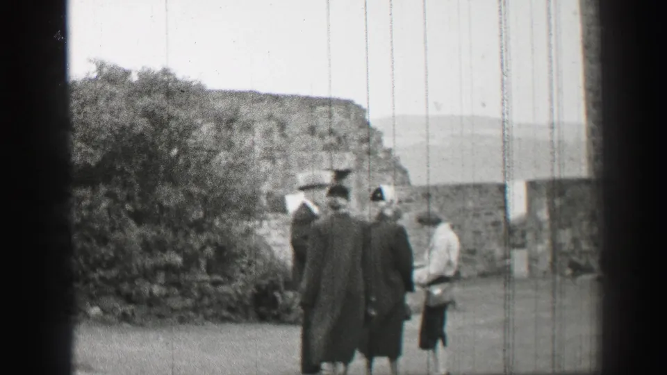 1952 United Kingdom 8mm frame showing a small group of formally dressed visitors standing near castle ruins and shrubbery.