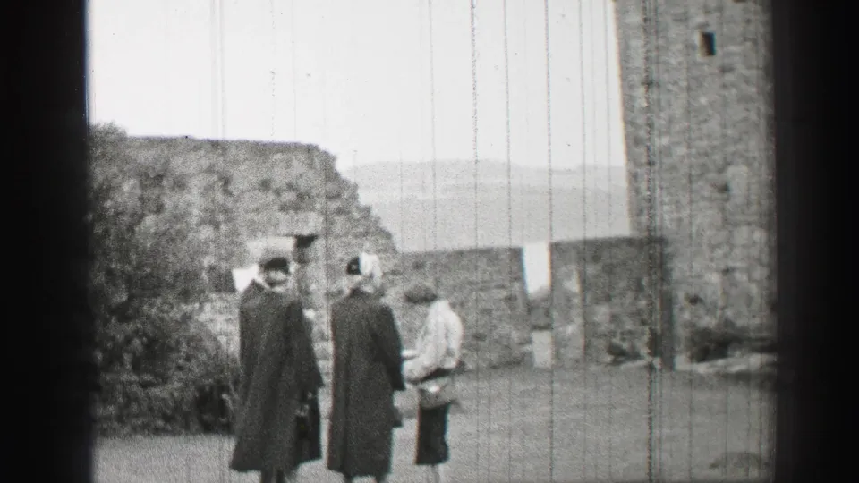 Women in mid-century coats and hats gather near stone walls during a United Kingdom castle courtyard visit.
