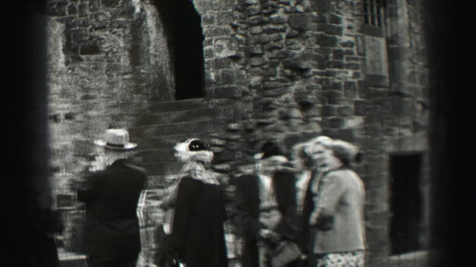 Visitors in coats and hats stand beside a weathered stone building, steps, and an arched doorway at a UK historic site.