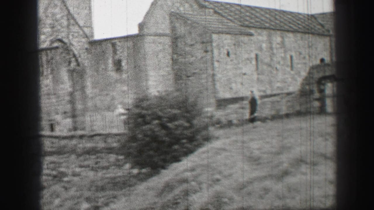Black-and-white 1952 United Kingdom 8mm frame with a stone structure, sloped grass, bush, and distant walking person.