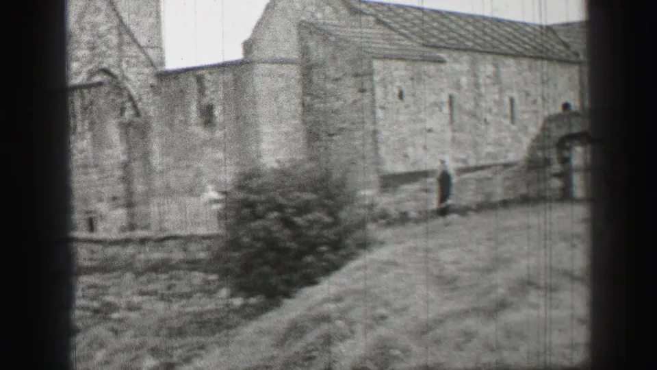 Black-and-white 1952 United Kingdom 8mm frame with a stone structure, sloped grass, bush, and distant walking person.