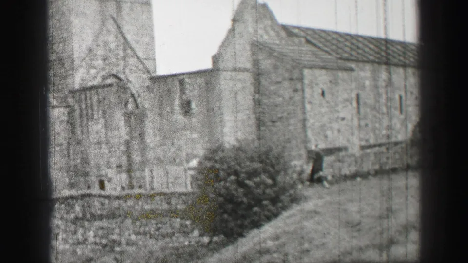 1952 United Kingdom home-movie frame showing a closer section of weathered stone architecture beside a bush and grass slope.