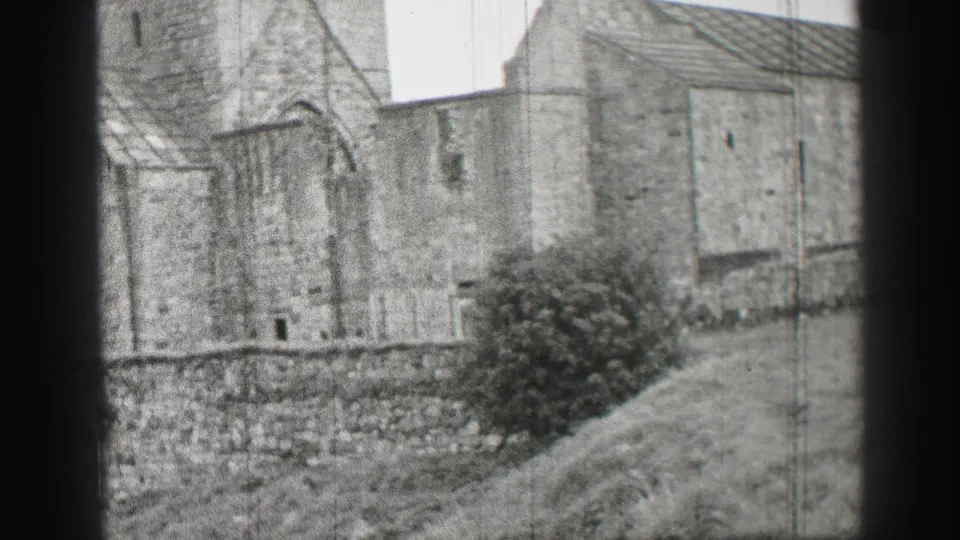 Black-and-white 8mm frame from the United Kingdom showing the stone building exterior, yard, bush, and a distant figure.