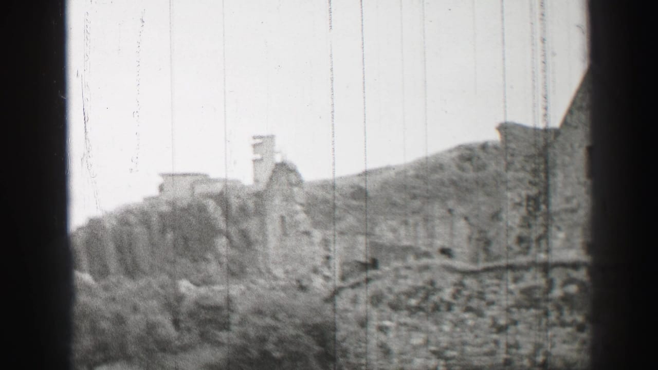 1952 United Kingdom 8mm frame showing weathered stone buildings, low walls, bushes, and a hillside in soft black-and-white focus.