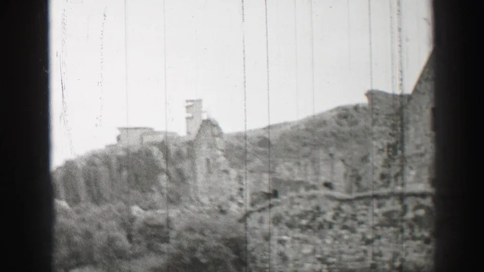1952 United Kingdom 8mm frame showing weathered stone buildings, low walls, bushes, and a hillside in soft black-and-white focus.