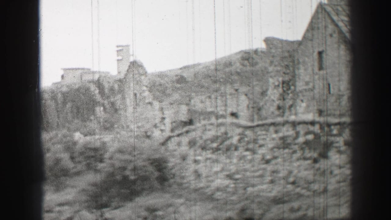 Old stone wall and roofed building beside overgrown ground in black and white.