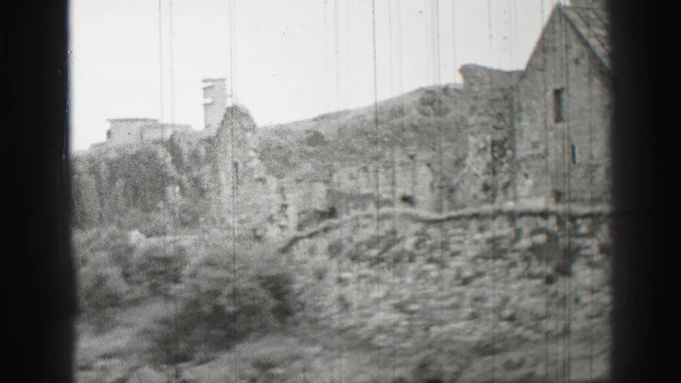 Black-and-white 8mm frame from the United Kingdom showing a stone wall, roofed ruin-like building, bushes, and rising ground behind.