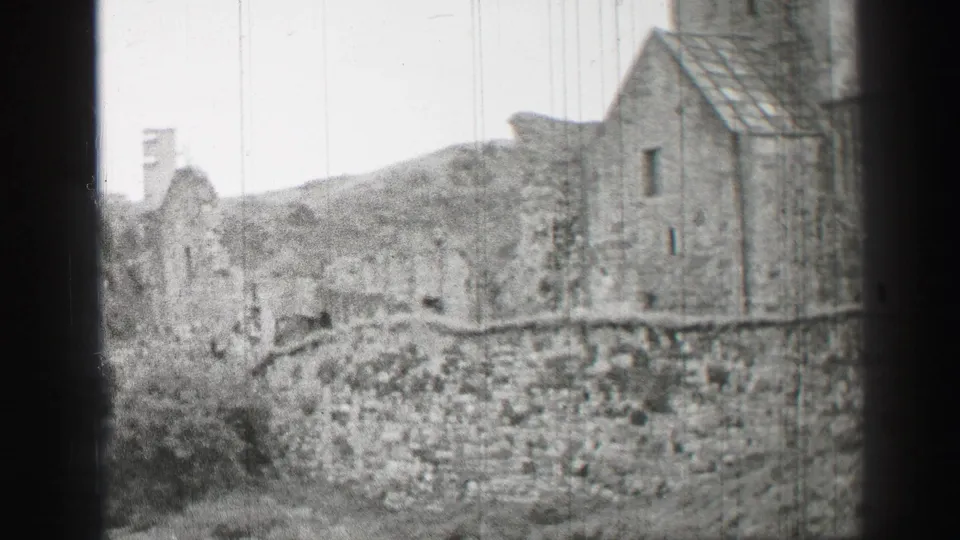 1952 United Kingdom home-movie frame showing a closer view of weathered masonry, small windows, bushes, and uneven sloped ground.