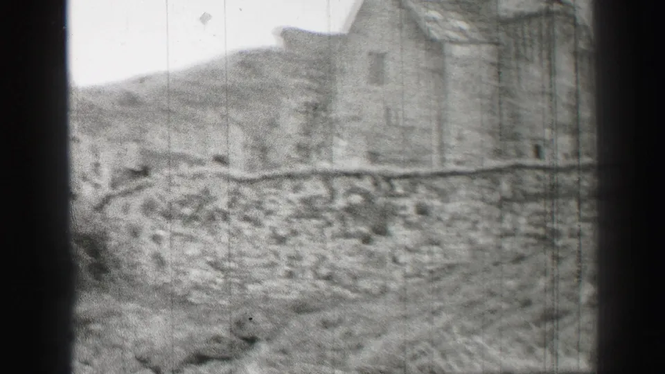 Black-and-white 8mm frame showing the edge of an old stone building, a low wall, brush, and hillside in the United Kingdom.