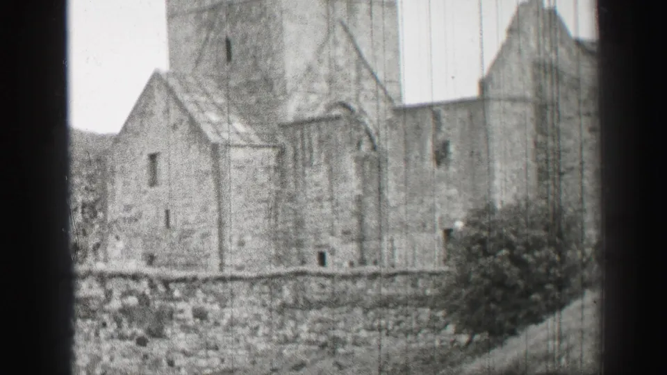 1952 United Kingdom 8mm view of weathered stone walls, a tower edge, small openings, and vegetation below the ruin.